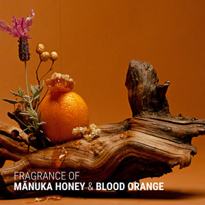 Decorative arrangement with an orange, lavender, and wooden piece on a brown background with text 'Fragrance of Manuka Honey & Blood Orange'.