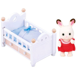 Small white crib with blue bedding and a rabbit figure in a red dress on a white background