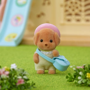 Small toy figure of a brown dog with a pink hat and blue bag on a green surface with flowers.