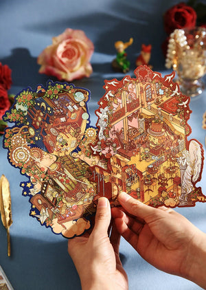A pair of hands holding a 3D papercraft pop-up model featuring an intricate Chinese-style landscape scene with golden pagodas and figures. The background is a soft blue fabric decorated with blurred roses and small figurines.