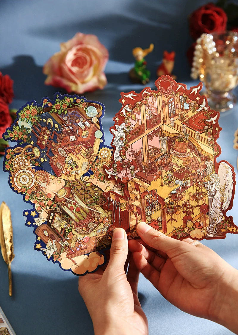 A pair of hands holding a 3D papercraft pop-up model featuring an intricate Chinese-style landscape scene with golden pagodas and figures. The background is a soft blue fabric decorated with blurred roses and small figurines.
