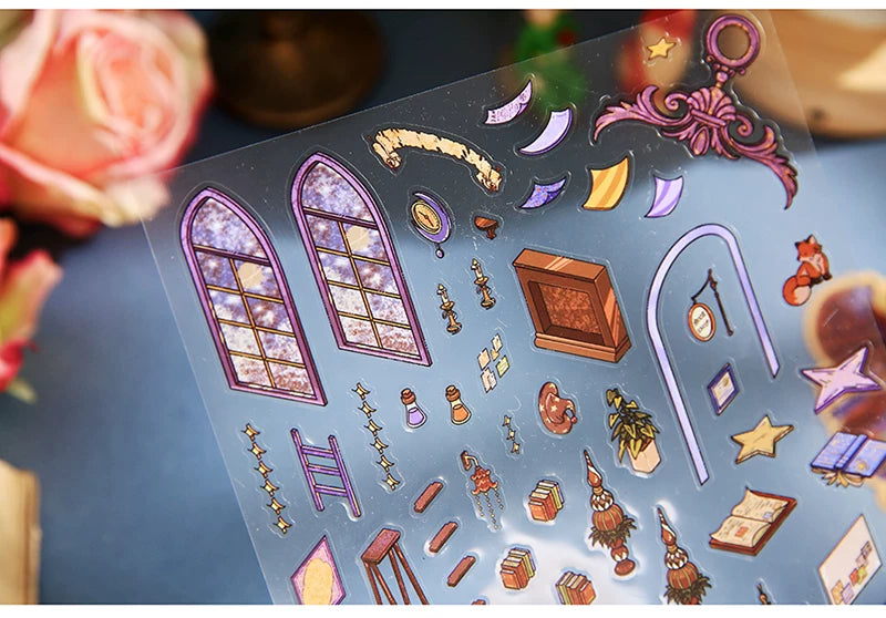 A clear sticker sheet featuring various whimsical, fantasy-themed illustrations including purple arched windows, crescent moons, stars, a small fireplace, books, and decorative objects, displayed on a blue surface with a blurred pink rose in the background.