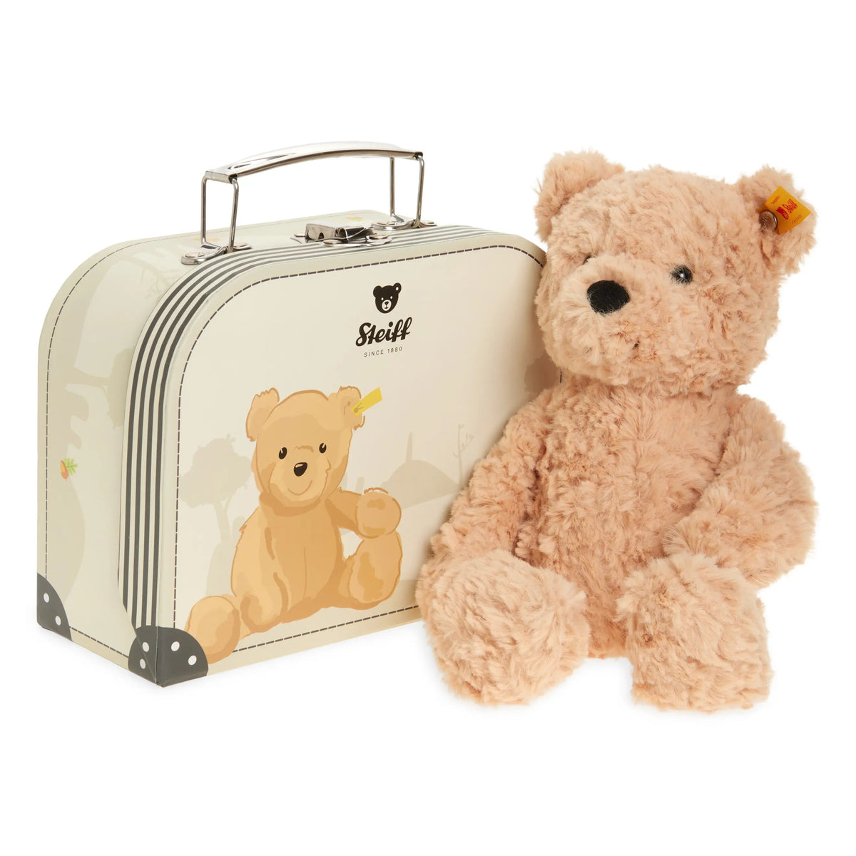 A Steiff teddy bear sitting next to its vintage cream-colored suitcase, featuring a printed teddy bear design and the Steiff logo on the suitcase, with a small orange tag visible on the bear's ear.