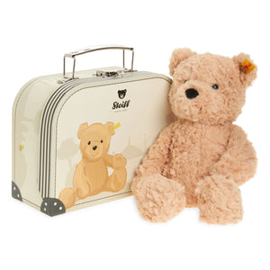 A Steiff teddy bear sitting next to its vintage cream-colored suitcase, featuring a printed teddy bear design and the Steiff logo on the suitcase, with a small orange tag visible on the bear's ear.