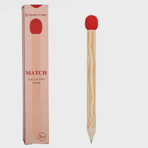 A gel ink pen shaped like a match, with a light wood-grain handle, a red circular top, and a silver tip, shown next to its pink packaging box on a light gray background.