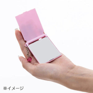 Hand holding a pink compact mirror with a heart charm on a white background