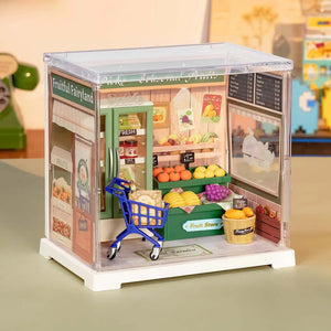 Miniature model of a fruit store with a shopping cart inside, on a neutral background.