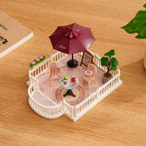 Miniature model of an outdoor cafe setup with chairs, table, and umbrella on a wooden surface.