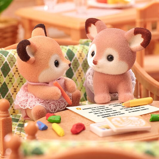 Two plush deer toys sitting at a table with crayons and paper.
