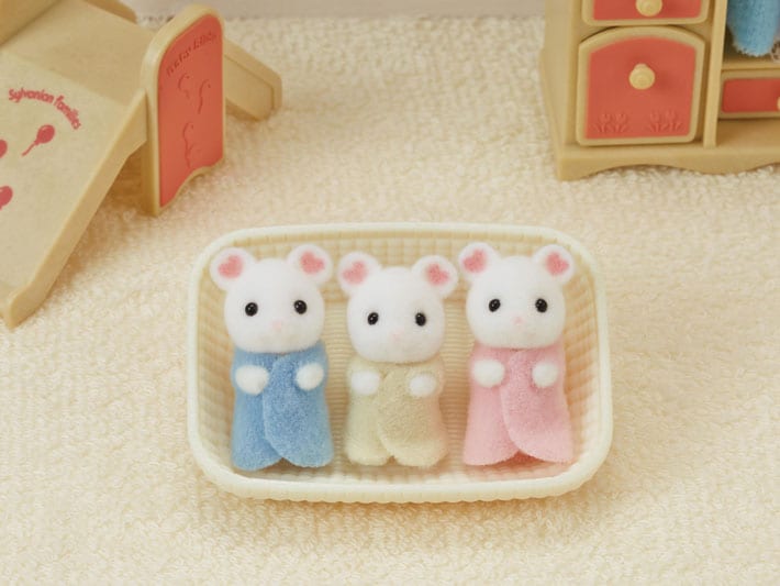 Three small animal figurines in pastel outfits on a textured surface with a toy house in the background.